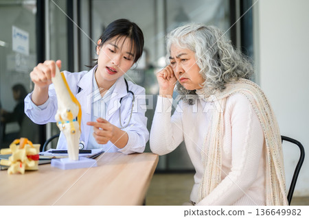 Asian woman doctor holding bone model talking to an old patient sitting at table in diagnosis room 136649982