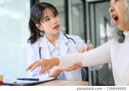 Asian woman doctor talking and holding arm of injured old patient sitting at table in diagnosis room Asian woman doctor talking and holding arm of injured old patient sitting at table in diagnosis room 136649989