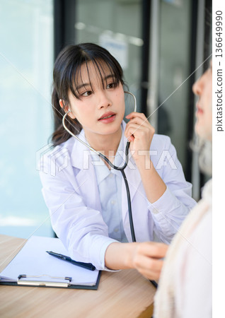 Asian woman doctor listening to patient's heartbeat with stethoscope sits at table in diagnosis room Asian woman doctor listening to patient's heartbeat with stethoscope sits at table in diagnosis room 136649990