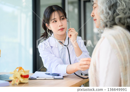 Asian woman doctor listening to heartbeat of patient with stethoscope sit at table in diagnosis room 136649991