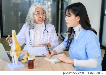Asian woman doctor taking note aside bone model while senior physician talking teaching with laptop. 136650012