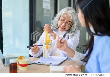 Old asian woman physician talking teaching on bone model with young doctor taking note at exam table Old asian woman physician talking teaching on bone model with young doctor taking note at exam table 136650016