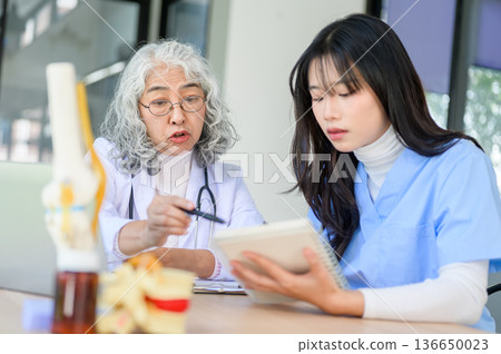 Old asian woman physician teaching and pointing at notebook of a young doctor sitting at exam table. 136650023