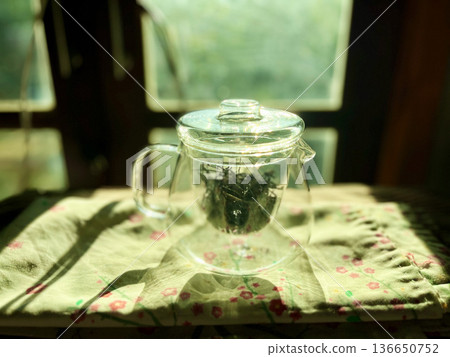 Glass teapot with green tea leaves on floral cloth in morning sunlight, minimalist zen lifestyle concept, Glass teapot with green tea leaves on floral tablecloth in morning sunlight 136650752