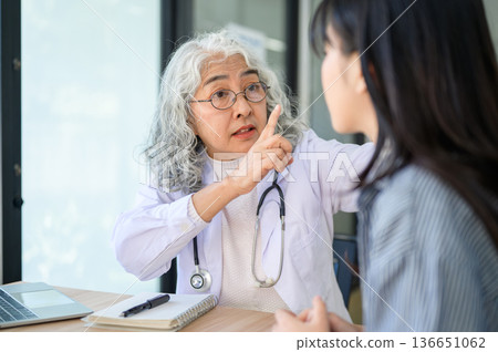 Old asian woman doctor warning or talking to young patient while sitting at table in diagnosis room. 136651062