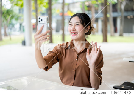 Asian woman student holding phone talking on video call while sitting at table in open air building. 136651420