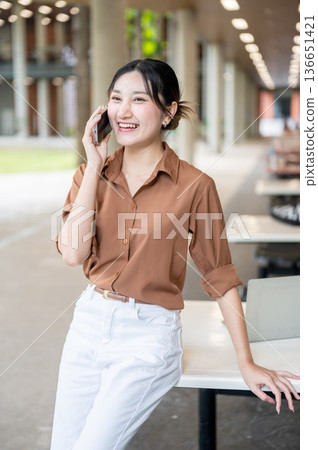 Asian woman student talking on phone call while standing leaning against table in open air building. Asian woman student talking on phone call while standing leaning against table in open air building. 136651421
