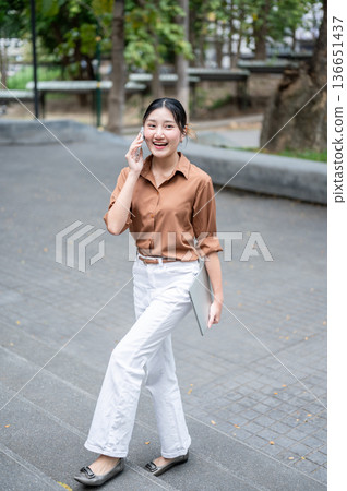 Smiling asian woman student talking on phone call carrying a laptop while walking on stairs in park. Smiling asian woman student talking on phone call carrying a laptop while walking on stairs in park. 136651437