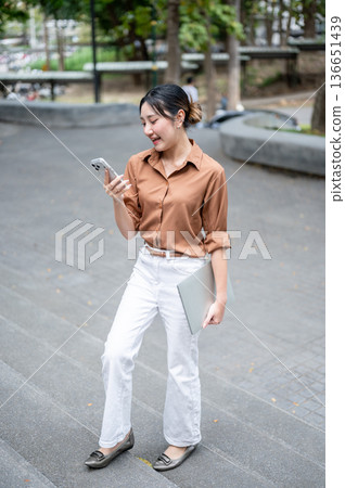 Asian woman student looking at phone and carrying a laptop while walking on concrete stairs in park. Asian woman student looking at phone and carrying a laptop while walking on concrete stairs in park. 136651439