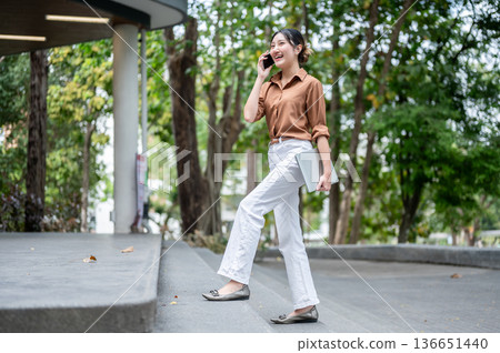 Full body shot of asian woman student talking on phone carrying laptop and walking on stairs in park Full body shot of asian woman student talking on phone carrying laptop and walking on stairs in park 136651440