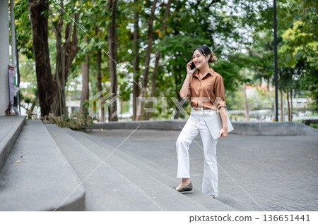 Happy asian woman student talking on phone call carrying laptop while walking on stairs in the park. Happy asian woman student talking on phone call carrying laptop while walking on stairs in the park. 136651441