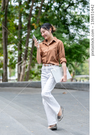 Happy asian woman student looking at phone and carrying laptop while walking on stairs in the park. Happy asian woman student looking at phone and carrying laptop while walking on stairs in the park. 136651442