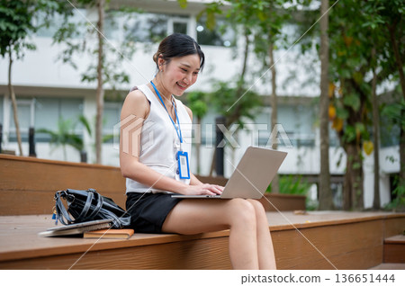 Asian office worker woman wearings lanyard working on laptop while sitting on wooden stairs in park. 136651444