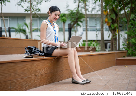 Asian office worker woman wearings lanyard typing on laptop while sitting on wooden stairs in park. 136651445