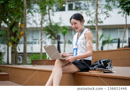 Asian office worker woman with lanyard watching video on laptop as sitting on wooden stairs in park. Asian office worker woman with lanyard watching video on laptop as sitting on wooden stairs in park. 136651446
