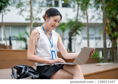 Asian office worker woman wearings lanyard looking at laptop while sitting on wooden stairs in park. Asian office worker woman wearings lanyard looking at laptop while sitting on wooden stairs in park. 136651447