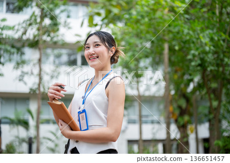 Asian woman office worker wearings lanyard holding pen and book while standing or walking in a park. Asian woman office worker wearings lanyard holding pen and book while standing or walking in a park. 136651457
