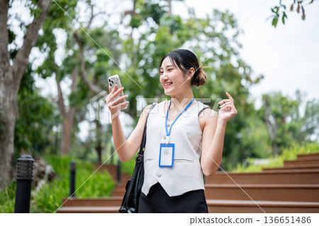 Asian woman office worker looking and smiling at phone while standing or walking on stairs in park. 136651486