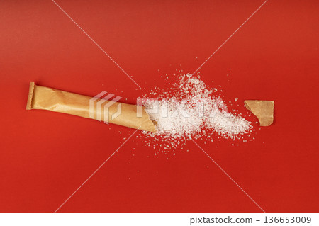 A torn craft paper sugar packet with white granulated sugar crystals scattered on a bright red surface. The composition shows a minimalist and conceptual view of sweetness or energy. Isolated A torn craft paper sugar packet with white granulated sugar crystals scattered on a bright red surface. The composition shows a minimalist and conceptual view of sweetness or energy. Isolated 136653009