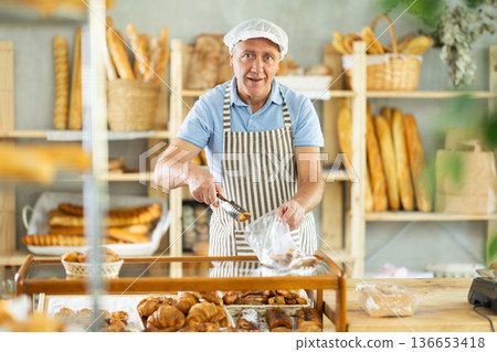 Senior man seller puts croissant in plastic disposable bag for buyer, packs goods. Senior man seller puts croissant in plastic disposable bag for buyer, packs goods. 136653418