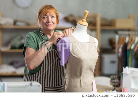 Mature female tailor sews new dress on a mannequin in sewing studio 136653580