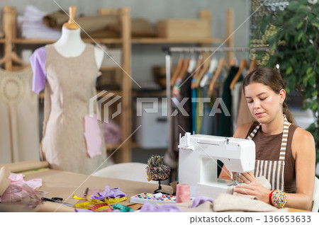 Female seamstress working on a machine against the background of a mannequin Female seamstress working on a machine against the background of a mannequin 136653633