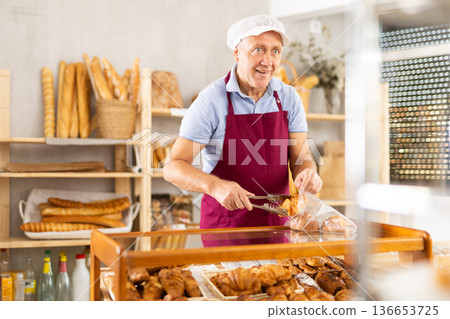 Senior man seller puts croissant in plastic disposable bag for buyer, packs goods. 136653725