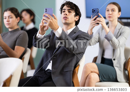 Positive young guy student sitting and listening lecture, taking photo or QR with mobil or tablet in lecture hall 136653803
