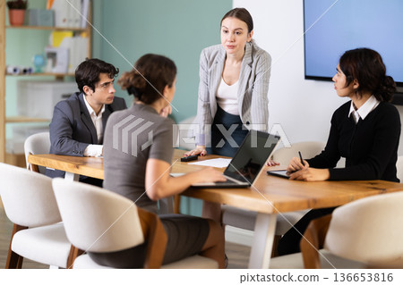 Confident young businesswoman standing near table where sitting colleagues talking and discussing ideas in office 136653816