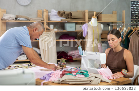 Girl employee sewing, stitch clothes, senior man pins paper pattern to fabric Girl employee sewing, stitch clothes, senior man pins paper pattern to fabric 136653828