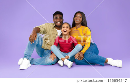 A family of three is sitting on a purple background. The father and mother are sitting beside their daughter. They are smiling and having fun. The daughter is laughing and is in the middle. A family of three is sitting on a purple background. The father and mother are sitting beside their daughter. They are smiling and having fun. The daughter is laughing and is in the middle. 136654247