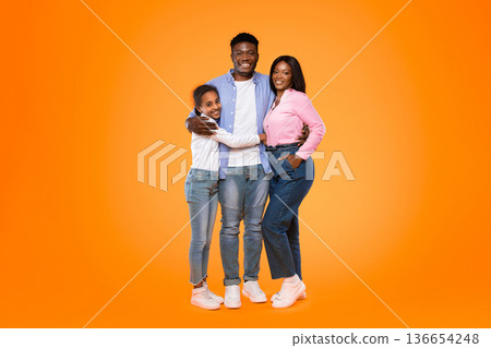 A man stands between a girl and a woman, all smiling and showing joy. They wear casual clothes. The bright orange background adds energy to the scene, capturing a fun family moment. A man stands between a girl and a woman, all smiling and showing joy. They wear casual clothes. The bright orange background adds energy to the scene, capturing a fun family moment. 136654248