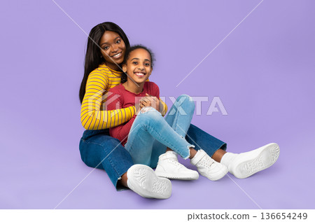 A mother and her daughter are sitting together on the floor against a purple background. They are smiling and enjoying their time together, showing affection and happiness. A mother and her daughter are sitting together on the floor against a purple background. They are smiling and enjoying their time together, showing affection and happiness. 136654249