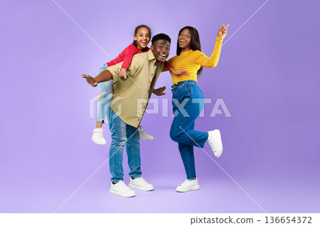 A man gives a piggyback ride to a smiling girl while a woman strikes a joyful pose next to them in front of a purple backdrop. They all display happiness and energy. 136654372