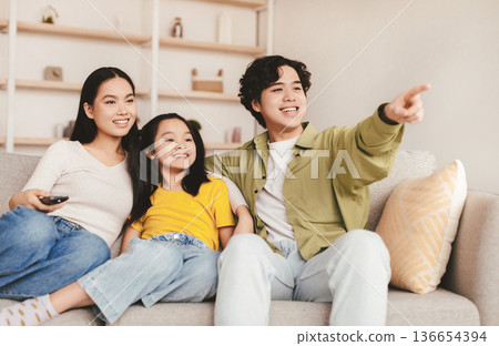 Smiling japanese young man shows on empty space with hand to wife and teen girl, sit on sofa in living room interior. Great offer, advertising, interesting movie at home together, facial expression 136654394