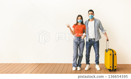 Young spouses wearing medical face masks standing with suitcase, woman pointing aside at free space, demonstrating place for your text or ad. Travelling during pandemic Young spouses wearing medical face masks standing with suitcase, woman pointing aside at free space, demonstrating place for your text or ad. Travelling during pandemic 136654395