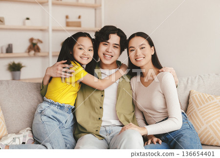 Glad smiling teen chinese girl, millennial family hugging on sofa and enjoy free time in minimalist living room interior. Couple with child spend weekend at home, relationship and covid-19 lockdown Glad smiling teen chinese girl, millennial family hugging on sofa and enjoy free time in minimalist living room interior. Couple with child spend weekend at home, relationship and covid-19 lockdown 136654401