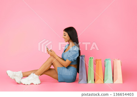 Shopping App. Laughing young woman using smartphone, sitting on floor with shopper bags, choosing and ordering clothes and presents isolated over yellow background, profile side view, free copy space Shopping App. Laughing young woman using smartphone, sitting on floor with shopper bags, choosing and ordering clothes and presents isolated over yellow background, profile side view, free copy space 136654403