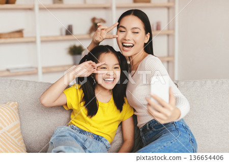 Satisfied teenage girl and young asian female have fun together, make selfie on smartphone in living room interior. Joy, video call, live stream, family blog and photo together at home during covid-19 Satisfied teenage girl and young asian female have fun together, make selfie on smartphone in living room interior. Joy, video call, live stream, family blog and photo together at home during covid-19 136654406