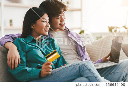 Happy young married Asian couple with laptop and credit card buying goods online, using modern pc for remote shopping at home, copy space. Internet banking, e-commerce, electronic money concept 136654408