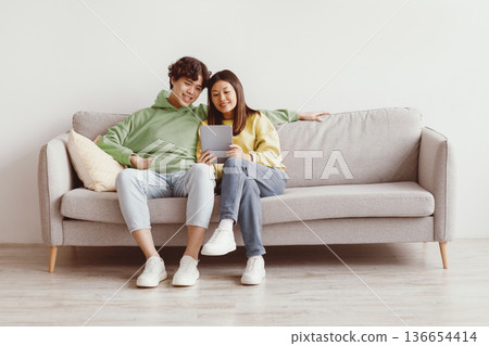 Happy Asian couple sitting on couch with digital tablet, looking at touch pad screen and smiling, using newest application at home, copy space. Millennial spouses browsing web, watching movie together Happy Asian couple sitting on couch with digital tablet, looking at touch pad screen and smiling, using newest application at home, copy space. Millennial spouses browsing web, watching movie together 136654414