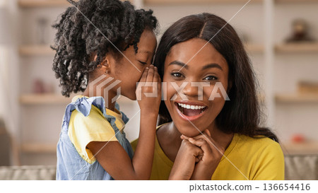 A young girl whispers to her mother while they sit in a bright living room. Both are smiling and enjoying their time together. The setting is warm and welcoming, with soft light. 136654416