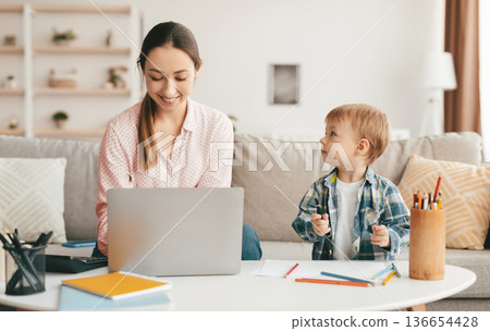 Adorable toddler boy drawing next to her mom working online on laptop in living room interior. Young mother sitting at table with computer, spending time with son and enjoying freelance work Adorable toddler boy drawing next to her mom working online on laptop in living room interior. Young mother sitting at table with computer, spending time with son and enjoying freelance work 136654428