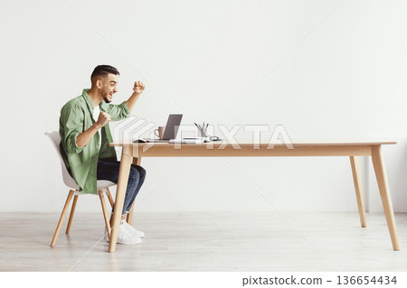 A man sits at a desk working on a laptop. He raises his arms in excitement, showing joy after achieving a goal. The office space is simple with a light-colored wall and wooden furniture. A man sits at a desk working on a laptop. He raises his arms in excitement, showing joy after achieving a goal. The office space is simple with a light-colored wall and wooden furniture. 136654434