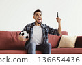 A man sits on a red couch with a soccer ball in one hand and a phone in the other. He looks excited and joyful while following a sports event on the phone. 136654436