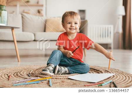Portrait of happy toddler boy sitting on carpet with colorful pencils and sheet of paper, drawing and playing at home, copy space. Adorable kid smiling 136654457