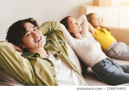Happy youngsters japanese girl, millennial mother relax, father with hand behind head look at camera on couch in minimalist room interior. Calm, peace, rest and silence at home, family enjoy free time Happy youngsters japanese girl, millennial mother relax, father with hand behind head look at camera on couch in minimalist room interior. Calm, peace, rest and silence at home, family enjoy free time 136654463