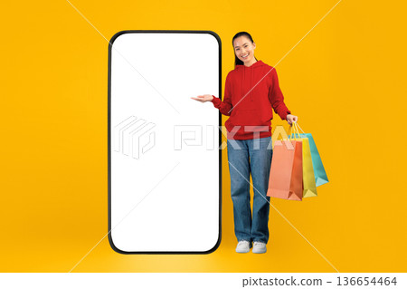 A woman stands next to a large blank phone screen, smiling and holding several colored shopping bags. The scene has a bright yellow background that adds energy to the moment. A woman stands next to a large blank phone screen, smiling and holding several colored shopping bags. The scene has a bright yellow background that adds energy to the moment. 136654464