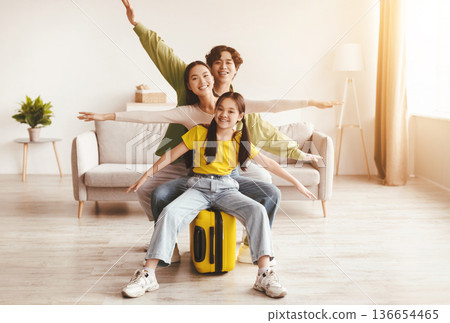 Happy excited korean young girl, mom and dad spread arms to sides like airplane, sit on yellow suitcase in living room. Preparation for travel, vacation with family, vacation after end of COVID-19 Happy excited korean young girl, mom and dad spread arms to sides like airplane, sit on yellow suitcase in living room. Preparation for travel, vacation with family, vacation after end of COVID-19 136654465