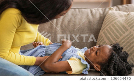 A mother plays with her young daughter on a couch inside their home. The daughter laughs and smiles while her mother interacts with her, creating a joyful moment. 136654470
